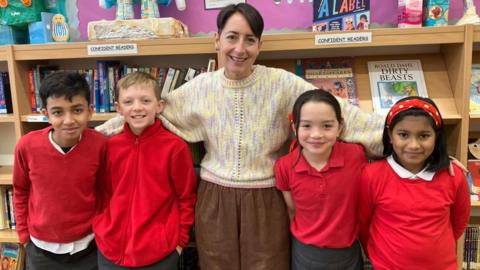 Deputy headteacher of Fishergate Primary School Dani Rees stands in the library with a mixed gender group of Fishergate Primary School pupils. Ms Rees wears a knitted jumper with purple, yellow and pink threads, and brown cord trousers. The pupils wear red jumpers or polo shirts.