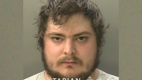 A mugshot of a man with lots of brown hair and facial hair. he is looking intensely, directly into the camera.