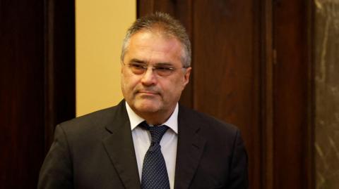A man with a black jacket and tie and dark hair and glasses stares at a camera