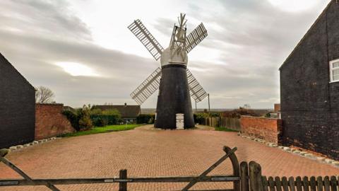 A black brick windmill with white wooden sales stands on a red-brick paved area. There are buildings on both sides and a fence in the foreground.