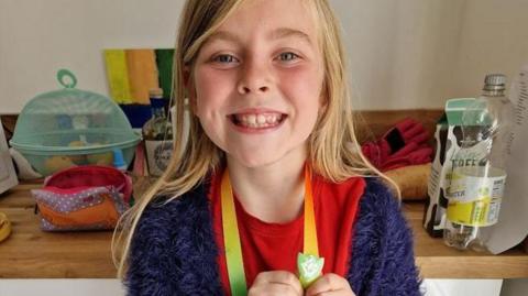 Matilda Pritchard holding her green Blue Peter badge for litter picking. She has long blonde hair and is wearing a blue cardigan and red t-shirt. She has a big smile and behind her is a wood kitchen work surface