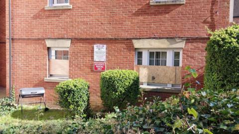 A brick-built house with its lower floor windows boarded up. The house is framed by green bushes in a garden area.