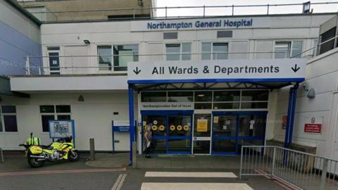 Entrance to hospital, a two-storey white building with white-framed windows. There are four blue doors, with a white one in the centre. A white sign with dark blue writing indicated "All Wards and Departments". There is a grey railing to the right and a motorcycle with rider in yellow hi-vis to the left.