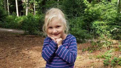 A young girl with blonde hair tied back stands in a wooded clearing. She is smiling and wearing a navy and pink striped jumper and with her hands by her chin.