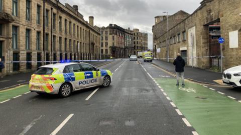 A police cordon on a two-way street. A police car is parked right next to the police tape, while a man in a black coat and pale jeans stands nearby. Industrial buildings flank the road on both sides.