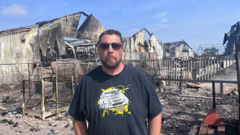 Adam Weaver stands with the fire wreckage behind him. He is wearing sunglasses and a black T-shirt with a Power Max logo on.
