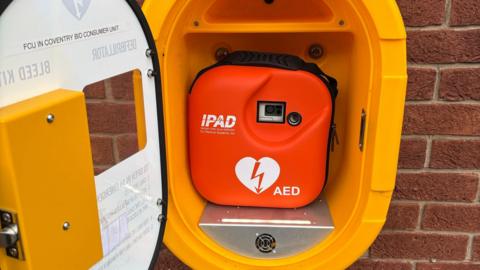 An orange box containing a defibrillator in a yellow cabinet attached to a brick wall.