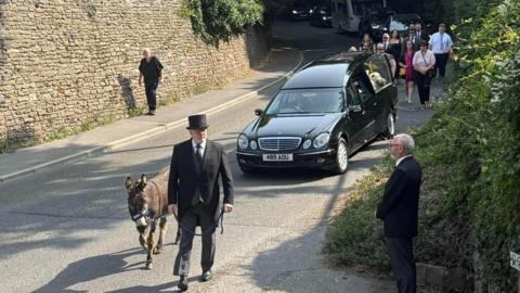 A black hearse being led by a man wearing a tuxedo suit and top hat, leading a brown donkey along the road.