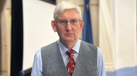 A grey haired man with glasses looks at the camera, wearing a blue shirt, grey waistcoat and red tie.