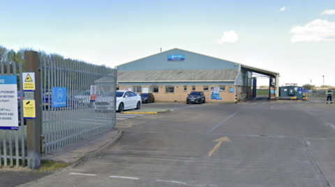 The two storey warehouse building is set behind a metal gate and a car park. The first floor is brick and the upper floor has a metal design on the exterior. A central blue sign reads: "Arriva". There is a structure on the right which looks like an arch, tall enough to hold a double-decker bus.