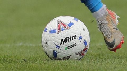 A National League football with 'HUFC' written on in pen being kicked by a grey boot and a blue sock