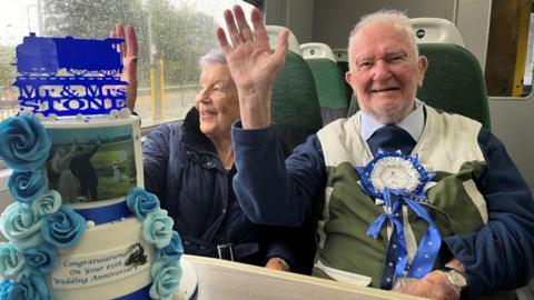 Close up of Margaret and George Stone waving while sat on a train with a cake infront of them