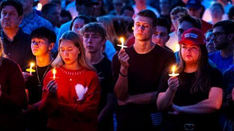 A candlelight vigil of students for the murdered right wing activist Charlie Kirk.
The scene is dimly lit, with the soft glow of candles illuminating the faces of those present. Each person holds a lit candle, and some clutch small crosses. One individual stands out wearing a red "Make America Great Again" hat. 
Visually, there is a contrast between the warm candlelight and the surrounding darkness. e expressions on the faces—quiet, reflective, and serious—underscore the gravity of the moment.