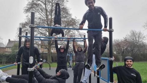 A group of people use the outdoor bar‑based fitness structure in a park. Some individuals are hanging from the bars, one is positioned upside down, and another is holding themselves above the bar. Others stand or lean on various parts of the structure. The scene takes place on a rubber‑surfaced exercise area with trees and grass in the background.