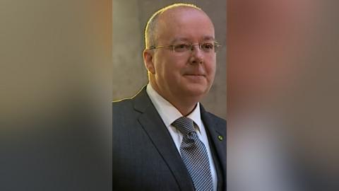 Former SNP chief executive Peter Murrell smiles as he looks off to the side. He is wearing glasses, a white shirt, dark grey blazer and a black and white tie.