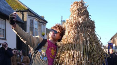 A man wearing a pink top and brown jacket and sunglasses is taking a selfie with the straw bear. 