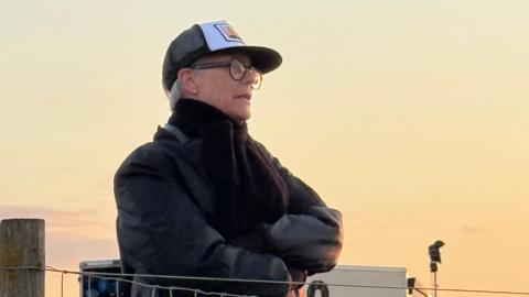 Jamie Lee Curtis behind a fence, wearing black jacket and cap