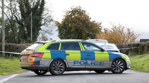 Yellow and blue police car at the scene of security alert in Keady, County Armagh. Car has a blue police logo on the side. There is police tape and trees and grass in the background.