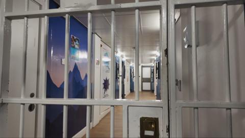 A photo of the bars of a police custody cell. Through the bars is a corridor which has pained artwork on the walls and messages