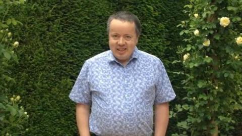 A man with grey hair smiling at the camera in front of green bushes. He is wearing a short sleeved blue patterned shirt.