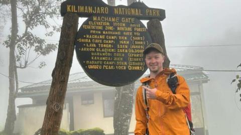 Freddie wears an orange raincoat and points to a sign that reads Kilimanjaro national park, that also includes timings to different points within the park.  It is very misty and wet.