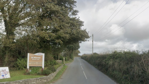 A view of a country road. A St Minver Holiday Park sign is on the left hand side of the road. Trees and bushes line either side of the road.