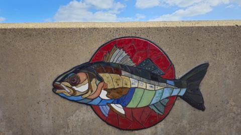 Mosaic of a fish on a sea wall - it is set with a red circle behind with the fish coloured in tiles of brown, green and blue.