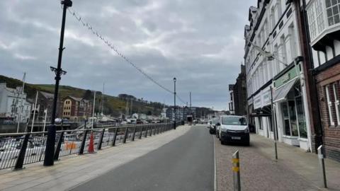 A street alongside a harbour marina, with a row of Victorian buildings that have a white facade on the right hand side of the street, and a railing and the boats in the the harbour on the left side. In the background are hills across the water.