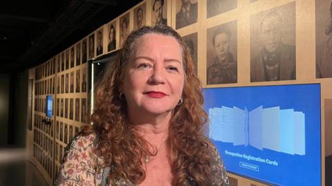 A woman with long brown curly hair standing in front of a wall with a number of portrait black and white photos on it