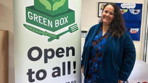 Melissa Trudgill, community engagement officer for Dartmoor Multi Academy Trust is wearing a dark blue patterned dress in front of a sign which reads: Green Box, Okehampton, Open to all!
