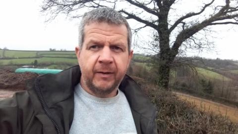 Jerry Saunders is standing outside. He has short grey hair and a short brown beard. He is wearing a grey jumper underneath and anorak. Behind him is a tree, hedging and the roof of a poultry shed in a field. It is a grey day.