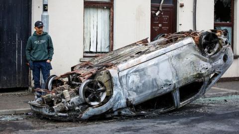 Man looking at burnt out overturned car