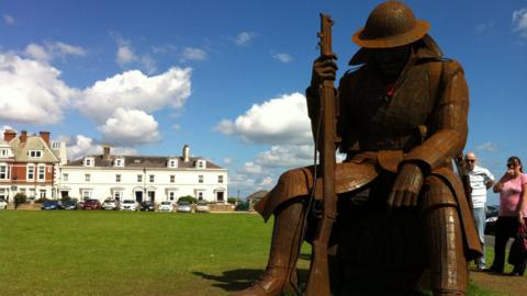 A statue of a World War One soldier sitting down and wearing a statue in Seaham. The large steel statue is brown in colour and holds a large rifle. A row of houses can be seen in the background.