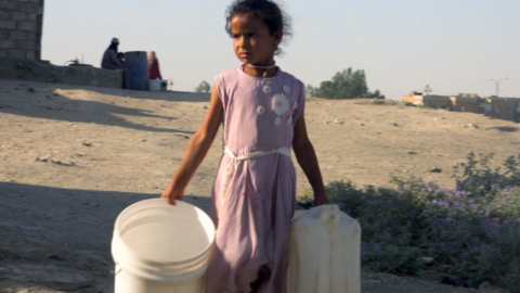 A young girl wearing a pink dress carries three empty plastic buckets and a cannister as she walks outdoors in Hassakeh