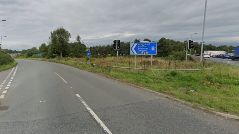 A blue motorway sign points to the M6 northbound junction 45. There is a road leading left and on the right is a grassy section where the signs and traffic lights stand. 