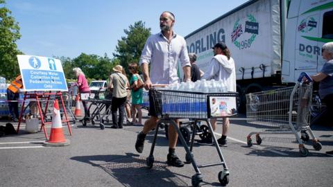 A man in a white shirt and shorts is pushing a trolley full of water bottles. People are queuing behind him and a lorry is in the background.