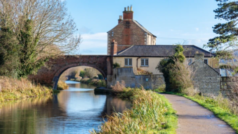 This photo has a river stream and a footpath. A bridge sits across the river with two buildings next to it. 