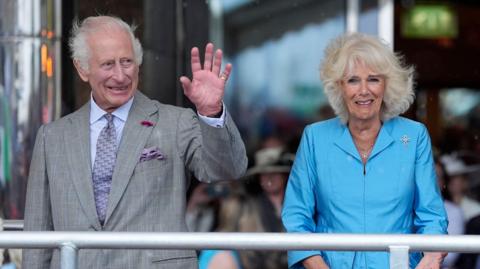 In the picture there is King Charles and Camilla. He is wearing a blue suit shirt with a grey blazer over the top. He has a purple tie and matching handkerchief, he is smiling and waving. Camilla is wearing a blue outfit, she is also smiling.