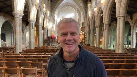 A man with white hair and wearing a blue gillet with a grey T-shirt underneath standing in a cathedral with wooden chairs either side of him and stone pillars.