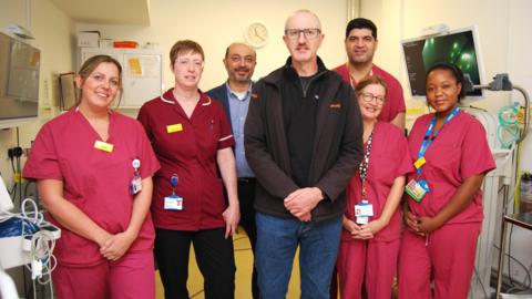 A man in a black quarter zip jumper and brown coat stands in the middle of the Colonoscopy team, dressed in pink hospital scrubs, at Kettering General Hospital.  