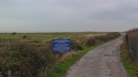 The entrance to Walney aerodrome. Grass field sits to the right with a blue sign that reads in white lettering - Walney Island Airfield. A concrete path runs along the grass verge to the right.