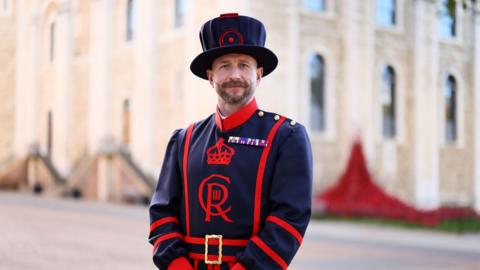 A man in Yeoman Warder uniform - a black cape with red outlines, black trousers, and a black cap.