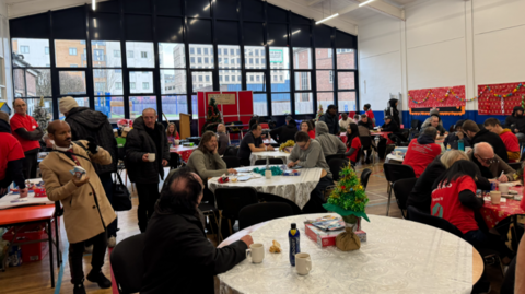 Dozens of people are pictured, with many of them sitting at round tables. A number of people are wearing red tops.