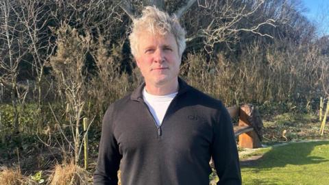 A man with fair hair, a white T shirt and a black sweater stands in front of a wooden bench and a line of trees.