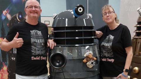 A man and a woman stood next to a shiny dark grey robot Dalek in matching t-shirts that say Dalek Dusty. 