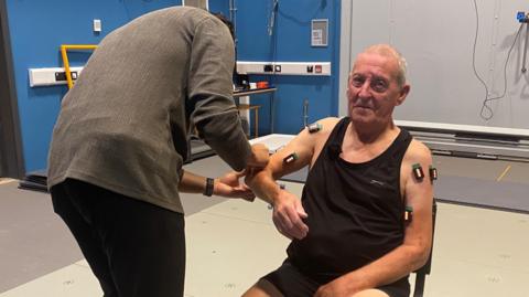 Bernie Kielty is sat down in a large room. He is wearing a black vest and has lots of different technology and equipment attached to his arms. There is a man in a grey top looking at his right arm.