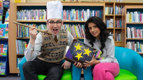Ben Shires and Konnie Huq sat in a library. Ben has a chef's hat on and is holding a wooden spoon. Konnie is holding a blue book with gold stars on the cover.