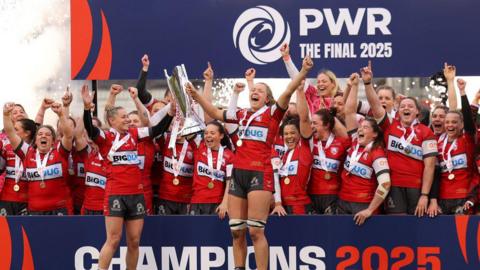 Gloucester-Hartpury players celebrate with the PWR trophy and a podium after winning the 2025 final