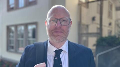 Deputy Rob Curgenven - A man with a bald head, a ginger and grey beard, wearing a blue suit, and a white shirt, with a blue and Guernsey flag logo tie. 