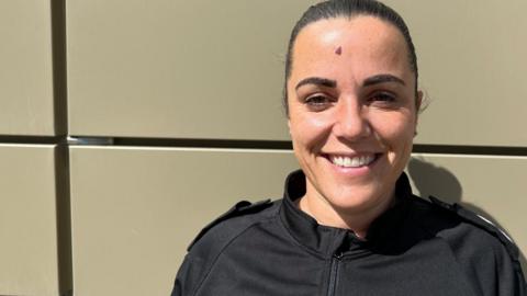 Inspector Jess Lawlor smiling at the camera, with dark hair in a ponytail, stood in front of a wall wearing a black top.